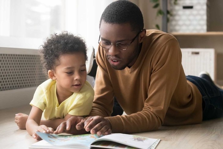 African American father in glasses and toddler son reading book together, lying on warm floor at home, black dad close up reading aloud fairy tale to adorable child, education, family weekend