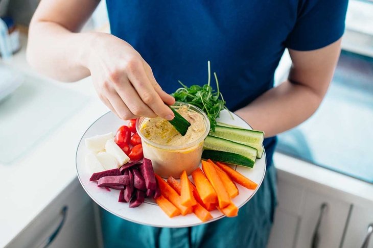 Top view close up man\'s hand dipping cucumber stick in hummus on the kitchen. Hummus served with raw vegetables on the plate. Healthy food lunch. Vegetarian and vegan food diet. Soft selective focus