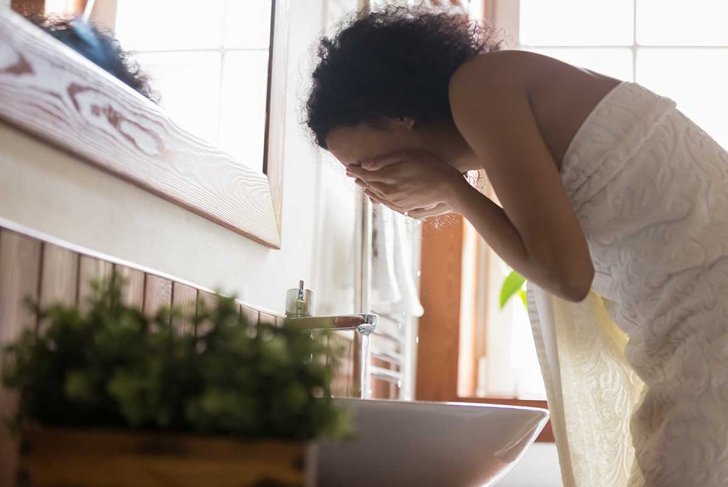 Side view young african american woman wrapped in towel after showering, bending over sink, washing off mask or peeling, cleaning her face with pure running water. Daily hygiene skincare routine.