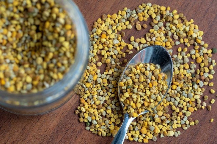 teaspoon full of bee pollen, on a table with scattered pollen granules, next to a vase, out of focus, of glass full of granules, dark wooden table background