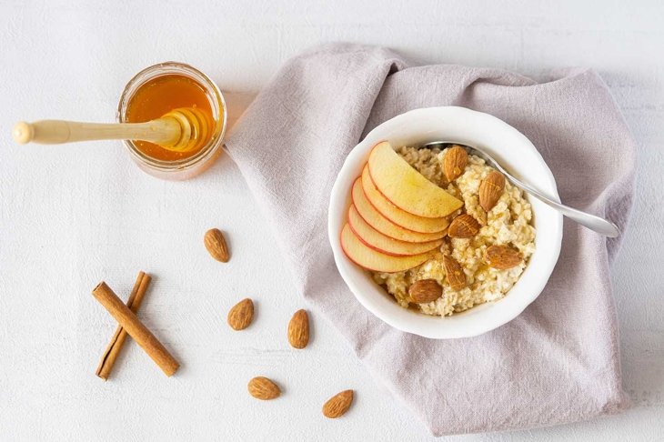 Oatmeal porridge with honey, apple, and almond nuts in a bowl served on grey napkin. There is jar of honey, cinnamon, and almond nuts. Vegan or vegetarian breakfast. Grey background, top view.