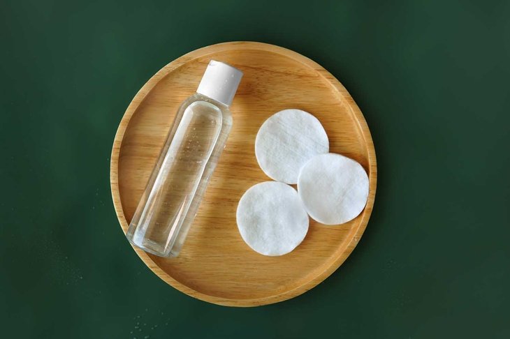 Top view First step routine Toner facial skincare product bottle with round organic cotton pad on bamboo wooden round plate with Tidewater Green solid color plain background with droplets