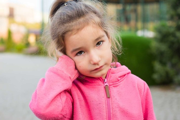 A little girl in a pink hoodie with a sad and tearful face is holding her ear. Ear pain, otitis media, swelling of the cheek, gums, toothache, children\'s surgery, otolaryngology. Children\'s medicine