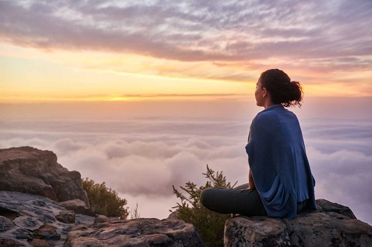 Rear view of a young woman sitting on a mountain top peacefully gazing at low-lying morning clouds and the pastel colours of a tranquil sunrise