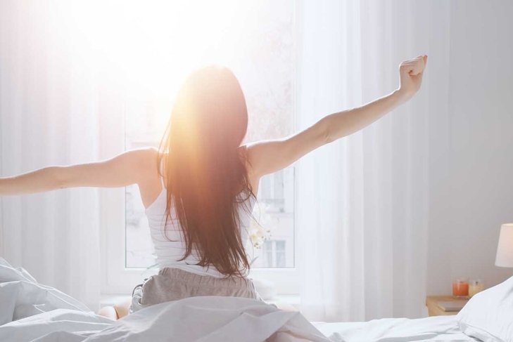 Beautiful Brunette is Waking up in the Morning, Stretches in the Bed, Sun Shines on Her From the Big Window. Happy Young Girl Greets New Day with Warm Sunlight Flare.