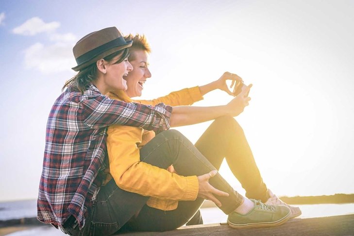Happy lesbian couple taking a selfie with mobile smart phone camera on the beach at sunset - Vignette edit - Homosexuality, diversity, vacation, travel, lgbt, technology concept