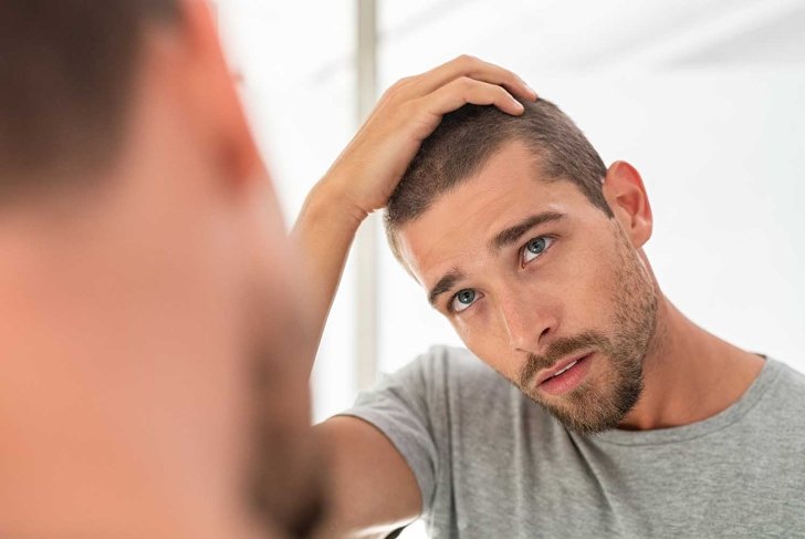 Young unshaven man looking at mirror in bathroom at home. Handsome guy looking at his face in mirror, checking hair and hairline. Man in pijamas concerned with hair loss.