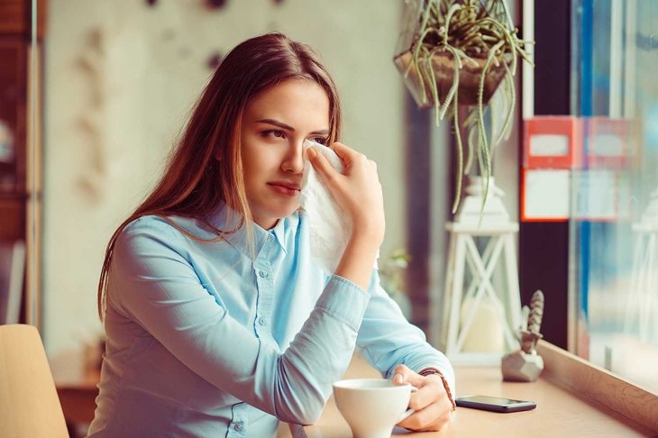 Misery, infelicity. Brunette woman girl about to cry wiping tears sneezing in a tissue drinking tea, coffee, hot beverage. Negative human emotion, face expression, reaction, body language