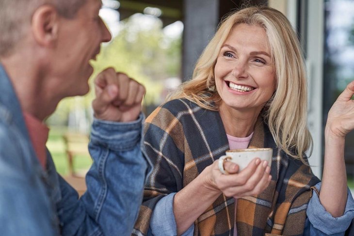 Cheerful mature lady is communicating with husband while relaxing with mugs of tea in green countryside