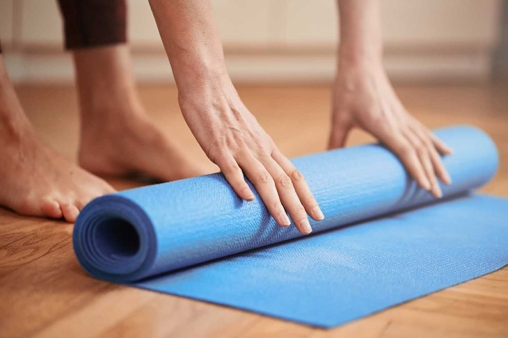Close up of mature woman folding blue yoga or fitness mat before or after working out at home in living room. Healthy life in covid-19 time lockdown. Focus on hand. Little bit of film grain